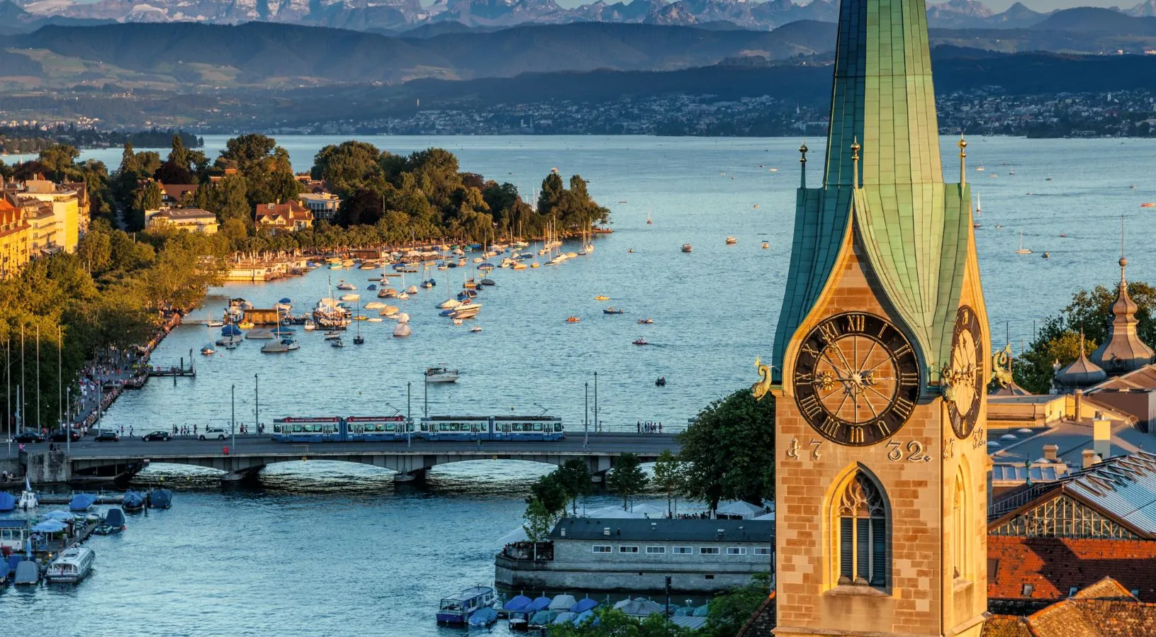 Scenic view of Lake Zurich with boats, the Fraumünster clock tower, and distant Alps at sunset, perfect for Switzerland honeymoon packages focused on city romance and lakeside charm.
