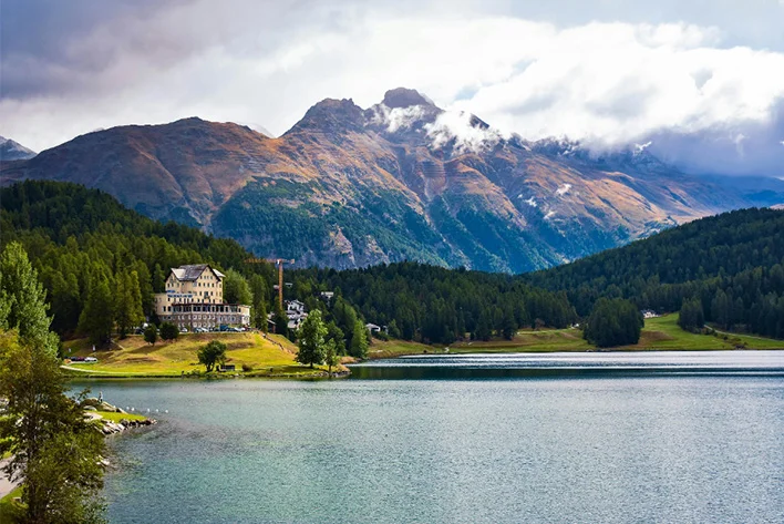 A panoramic view of Saint Moritz Lake, Switzerland with a building and mountains in the background.