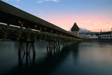 Chapel Bridge in Lucerne, Switzerland, at sunset, with the sky painted in warm hues of orange and pink.