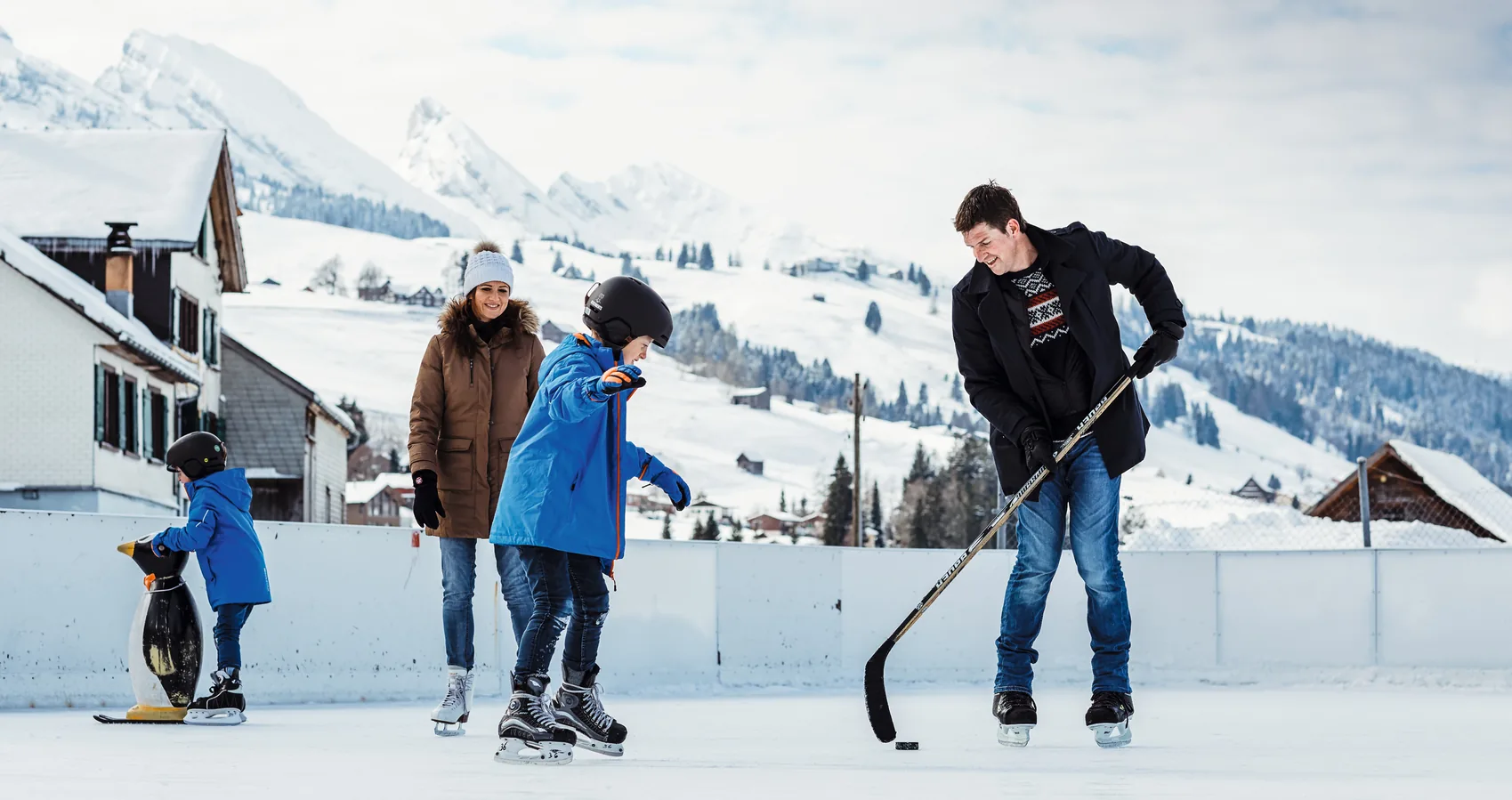 Family of four ice skating on an outdoor rink in Wildhaus with snow-covered Toggenburg peaks behind
