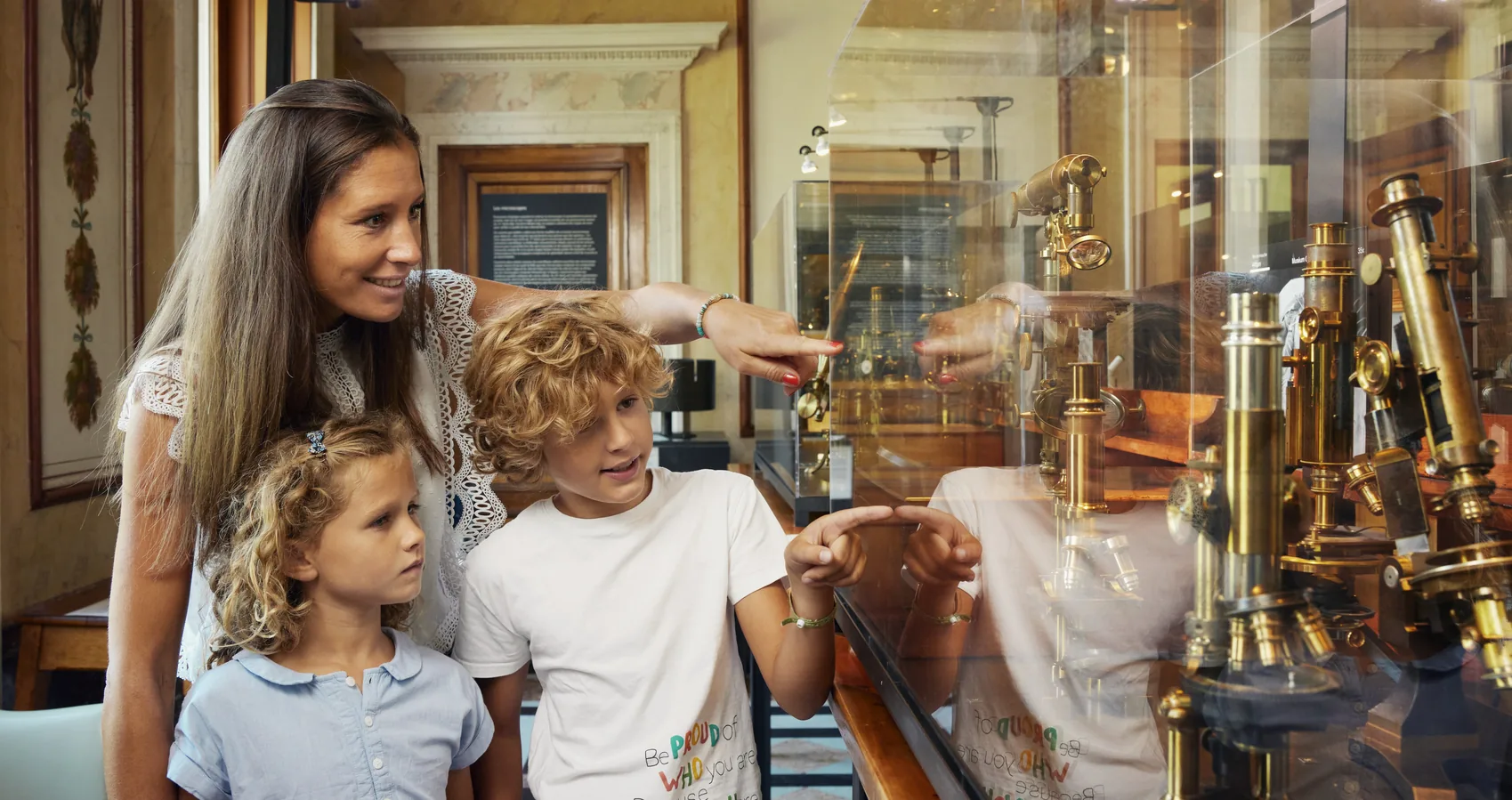 Mother and two children pointing at antique brass microscopes in a display case at a museum in Geneva