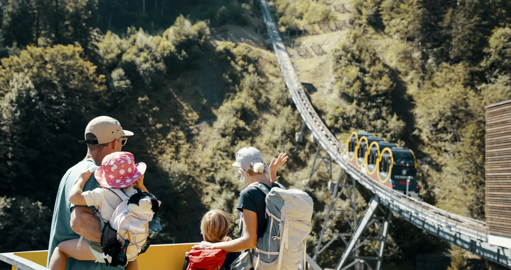 Family watching the Stoos funicular climb the steep mountainside from the valley station viewing platform