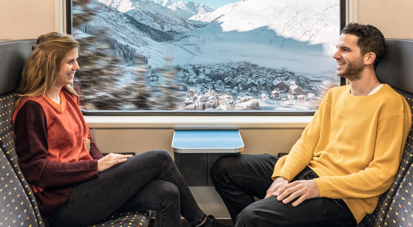 Two travellers chatting by a large train window with snowy mountains outside, illustrating how to use the Swiss Travel Pass on Swiss trains