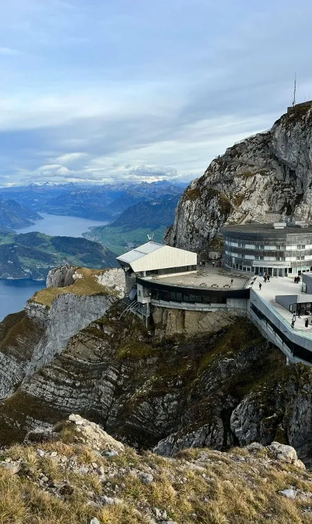 A breathtaking view of the Pilatus Mountain station, surrounded by rugged peaks and lush green valleys, with visitors enjoying the panoramic landscape and clear blue skies.