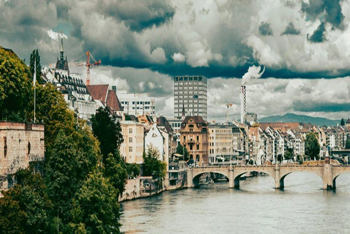 Scenic view of the Rhine River in Basel, Switzerland, featuring the tranquil waters flowing past historic buildings, vibrant waterfront promenades, and lush greenery along the riverbanks.