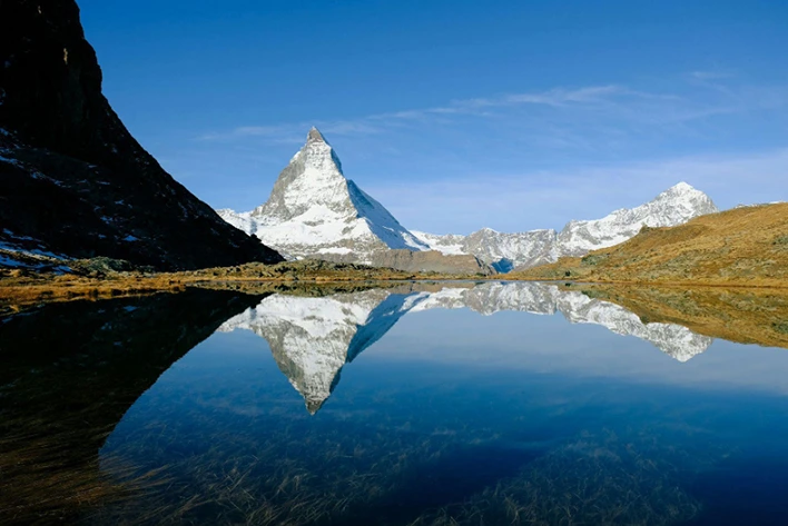 Serene view of Riffelsee in Zermatt, Switzerland, featuring the iconic reflection of the Matterhorn mountain in the tranquil waters of the lake, surrounded by picturesque alpine scenery and lush greenery.