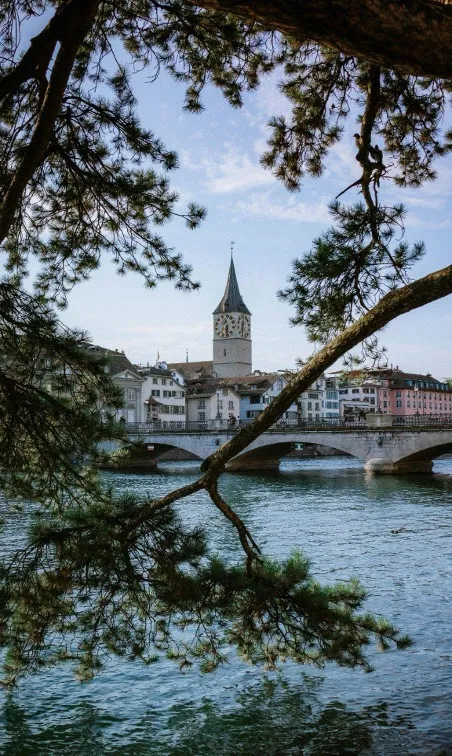 A picturesque view of St. Peter's Church in Zurich, showcasing its distinctive clock tower with the largest clock face in Switzerland, surrounded by charming historic buildings and a clear blue sky.