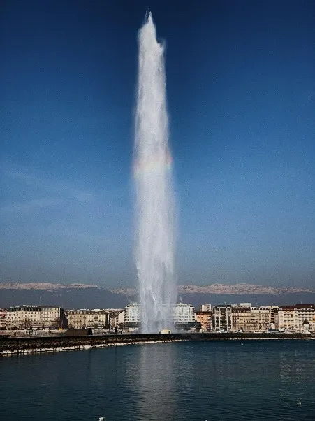 A captivating view of the Jet d'Eau, Geneva's famous water fountain, shooting high into the sky against a backdrop of the sparkling Lake Geneva and surrounding mountains, with blue skies and fluffy clouds enhancing the scene.